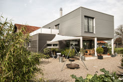 Modernes Einfamilienhaus mit Metallfassade, grosser Terrasse und Gartenbereich an sonnigem Tag.