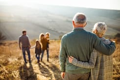 Seniorenpaar blickt über Landschaft auf Familie mit Kindern im Gegenlicht, Symbol für Generationen und Natur.