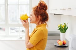 Junge Frau in gelbem Shirt spielt mit gelber Sparschweinfigur in heller Küche mit frischen Tulpen und Obst.